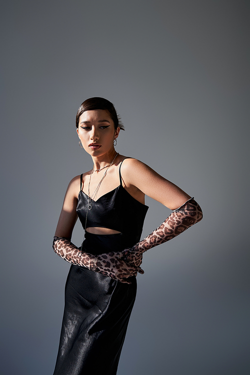 young and fashionable asian woman with bold makeup, brunette hair, in black strap dress and animal print gloves standing with hands on hip on dark grey background, spring fashion concept