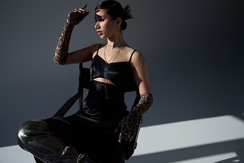 youthful syle, spring fashion, young asian woman in black elegant dress and animal print gloves sitting on chair in lighting on dark grey background, generation z, silver necklaces