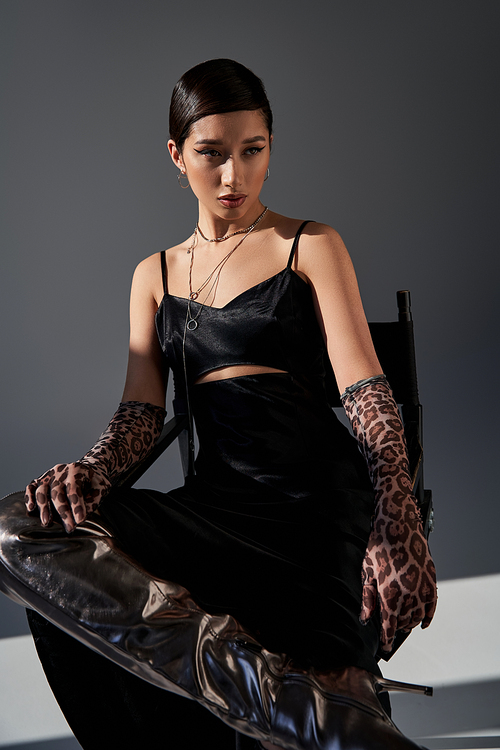 spring fashion concept, young asian woman with bold makeup, brunette hair, black strap dress, animal print gloves and silver boot sitting on chair on grey background with lighting, generation z