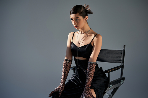 young asian fashion model with brunette hair posing on chair in trendy spring outfit, black strap dress, animal print gloves, silver necklaces on grey background with lighting, generation z