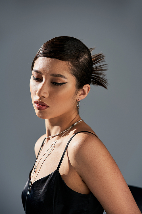 portrait of appealing asian woman with brunette hair, bold makeup, trendy hairstyle, in black strap dress and silver accessories posing in lighting on grey background, spring fashion concept