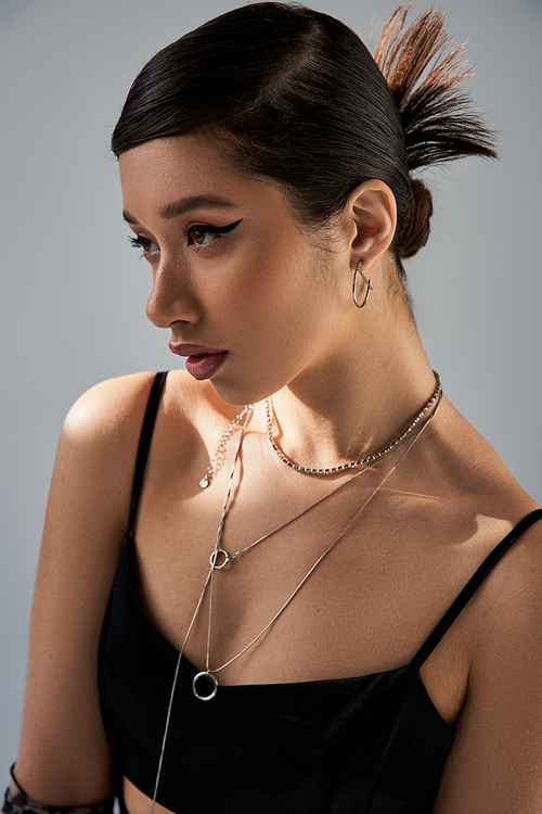 portrait of brunette asian woman with bold makeup, trendy hairstyle and expressive gaze, in black strap dress and silver necklaces looking away on grey background with lighting, trendy spring concept