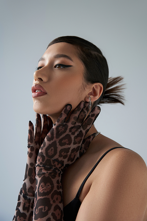 youthful style, spring fashion, young asian woman with brunette hair and bold makeup, wearing black dress and animal print gloves, touching neck, expressive, on grey background