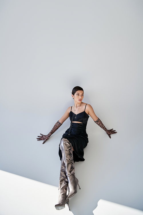 spring fashion photography, high angle view of elegant asian woman sitting in black elegant strap dress, animal print gloves and silver boots on grey background with lighting