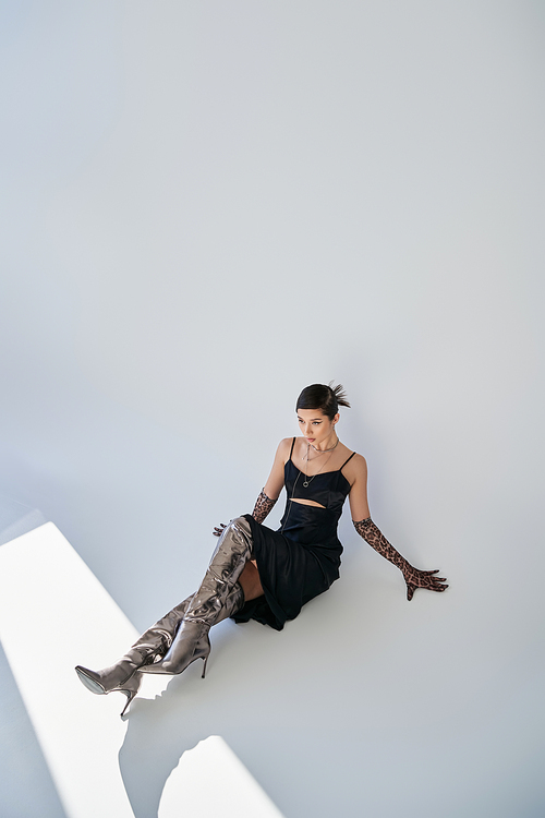 high angle view of asian woman with brunette hair, in black and elegant dress, silver boots and animal print gloves sitting on grey background, generation z, spring fashion photography