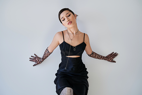 stylish asian woman with closed eyes and bold makeup sitting in black strap dress, animal print gloves and silver accessories on grey background, spring fashion photography