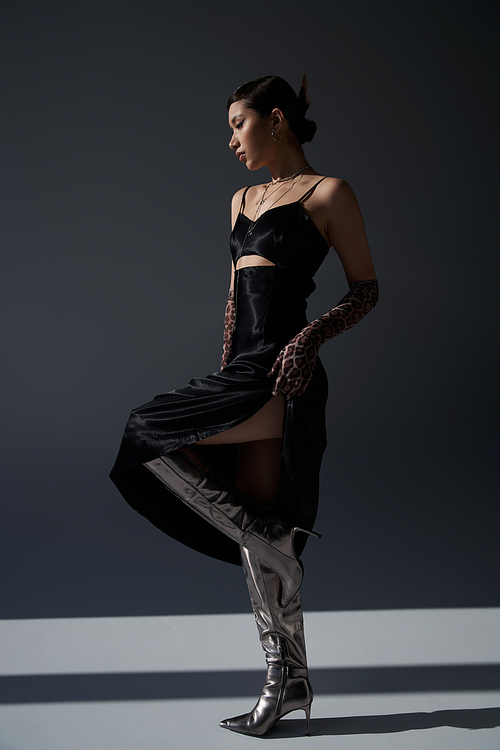 full length of graceful asian woman in black elegant dress, silver boots and animal print gloves standing on dark grey background with lighting, stylish spring, fashion shoot