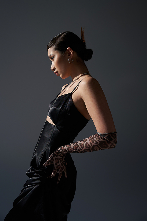 side view of young and pretty asian woman with trendy hairstyle posing with hand on hip in black strap dress and animal print glove on dark grey background, fashionable spring concept