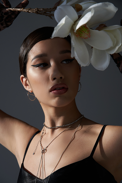 portrait of expressive asian woman in silver necklaces and black strap dress, with brunette hair and bold makeup holding white orchid on dark grey background, fashionable spring concept