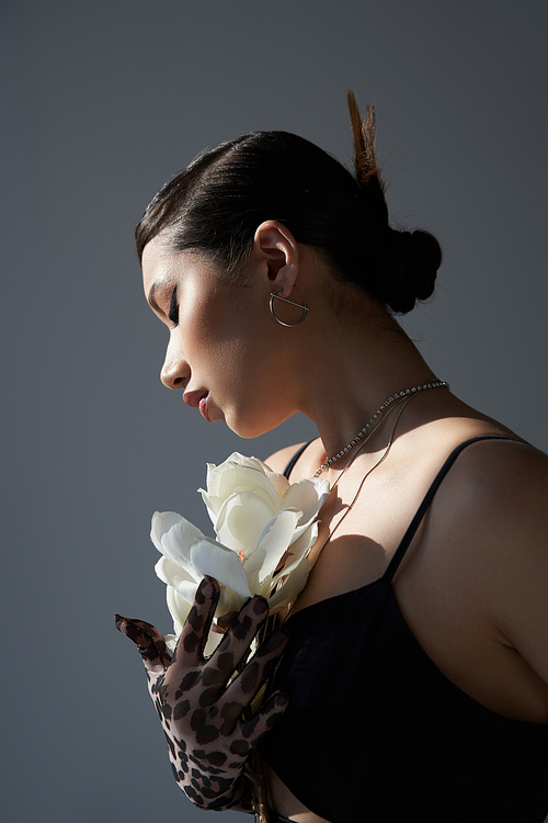 appealing asian woman with brunette hair, bold makeup and silver accessories, in black dress and animal print glove holding white orchid on dark grey background, stylish spring concept