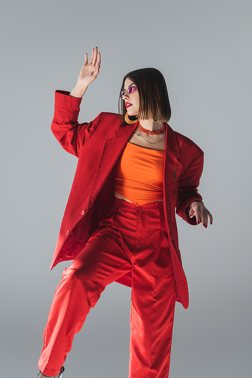 lady in red, young brunette woman with short hair posing in pink sunglasses and red suit and gesturing on grey background, generation z, trendy outfit, fashionable model, professional attire