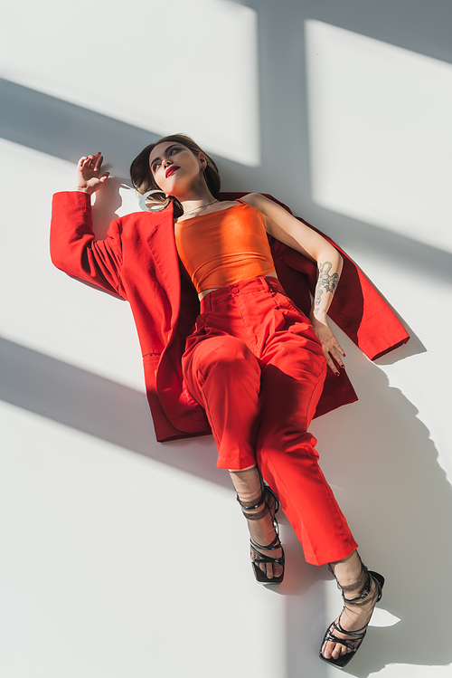 top view, young tattooed woman with short hair lying in red suit on grey background, generation z, fashionable model, professional attire, corporate fashion, heeled shoes, lady in red