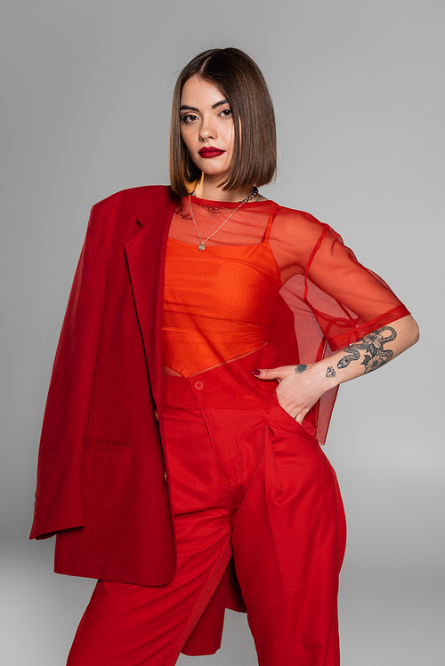 stylish suit, bold makeup, young tattooed woman with short hair holding red blazer on grey background, generation z, trendy outfit,  professional attire, executive style