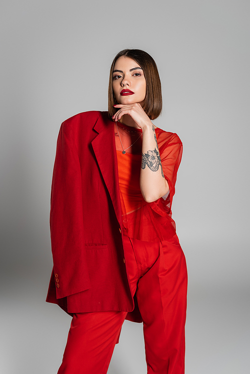 trendy outfit, bold makeup, tattooed woman with short hair holding red blazer on grey background, generation z, professional attire, executive style, young professional