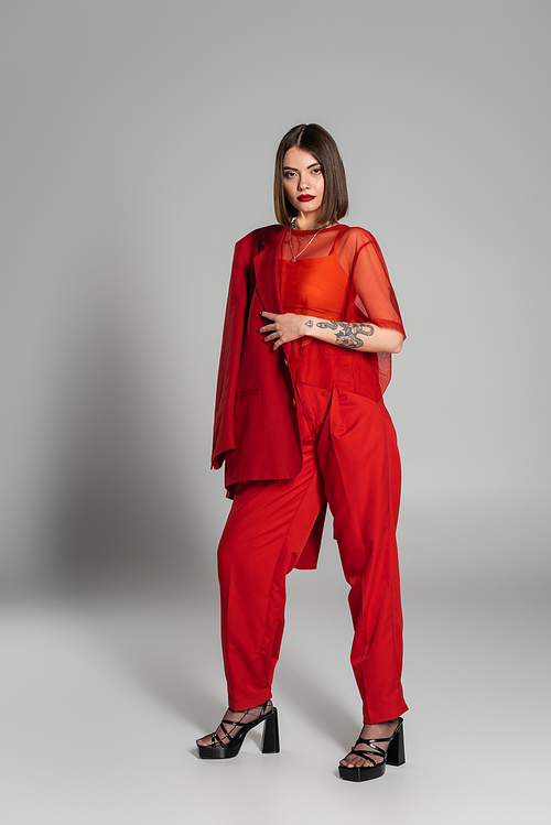 full length, trendy outfit, bold makeup, young tattooed woman with short hair holding red blazer on grey background, generation z, modern style, professional attire, executive style