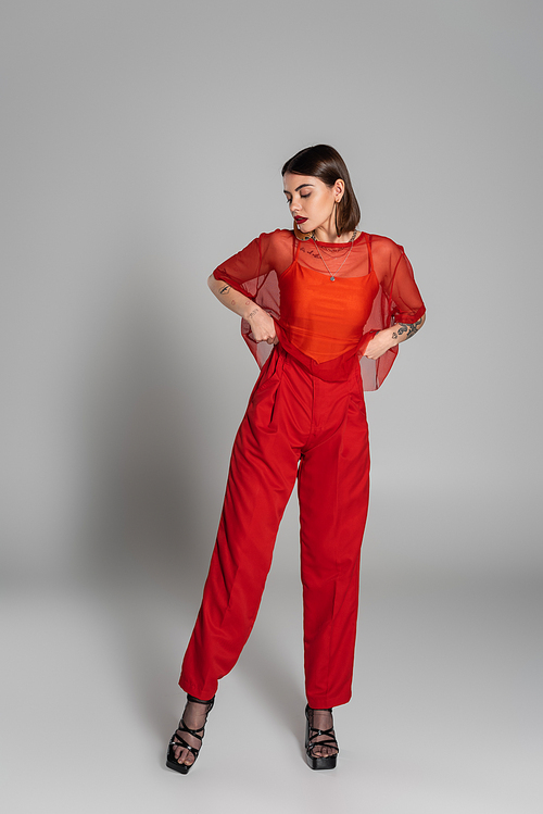 red outfit, tattooed and brunette woman with short hair and nose piercing posing in transparent blouse and pants on grey background, modern style, generation z, fashion photography, full length