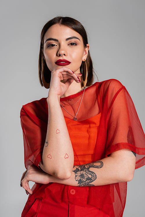fashion look, red outfit, tattooed and brunette woman with short hair and nose piercing posing in transparent blouse on grey background, modern style, generation z, portrait
