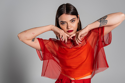 generation z, portrait of tattooed and brunette young woman with short hair and nose piercing posing in transparent blouse on grey background, modern style, fashion look, red outfit