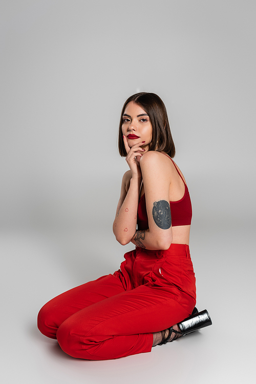 young model in red outfit, tattooed woman with short hair and nose piercing posing in red crop top and pants while sitting on grey background, modern style, generation z, fashion trend