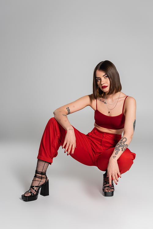 full length of young model in red outfit, tattooed woman with short hair and nose piercing posing in red crop top and pants while sitting on grey background, modern style, generation z, fashion trend