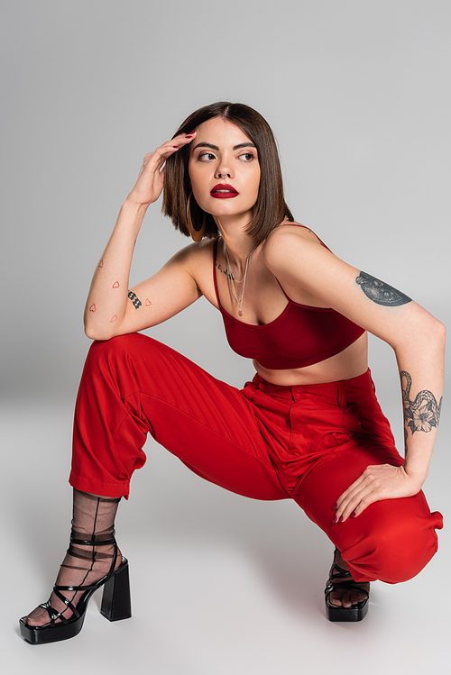 fashion trend, young model in red outfit, tattooed woman with short hair and nose piercing posing in red crop top and pants while sitting on grey background, modern style, generation z, full length