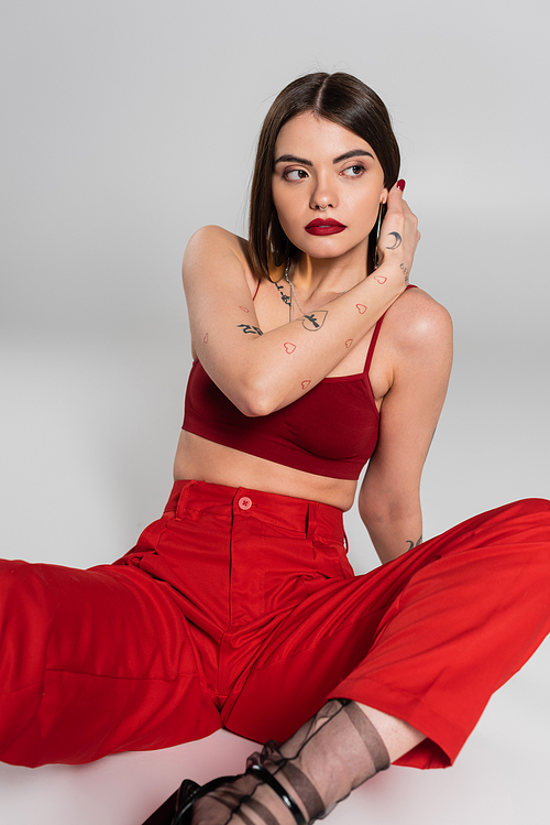 fashion trend, chic style, young model in red outfit, tattooed woman nose piercing posing in red crop top and pants on grey background, generation z, adjusting shirt hair