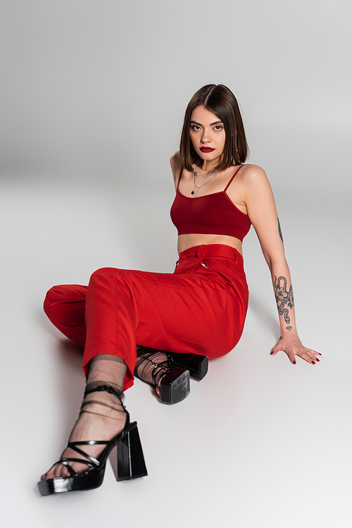 chic style, fashionable outfit, young model in red outfit, tattooed woman with short hair and nose piercing posing in red crop top and pants on grey background, generation z, full length