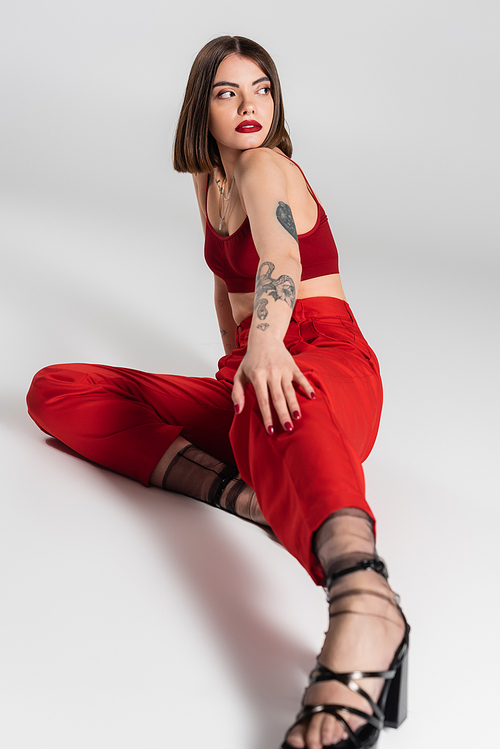 fashionable outfit, young model in red outfit, confident and tattooed woman with short hair and nose piercing posing in red crop top and pants on grey background, generation z, full length
