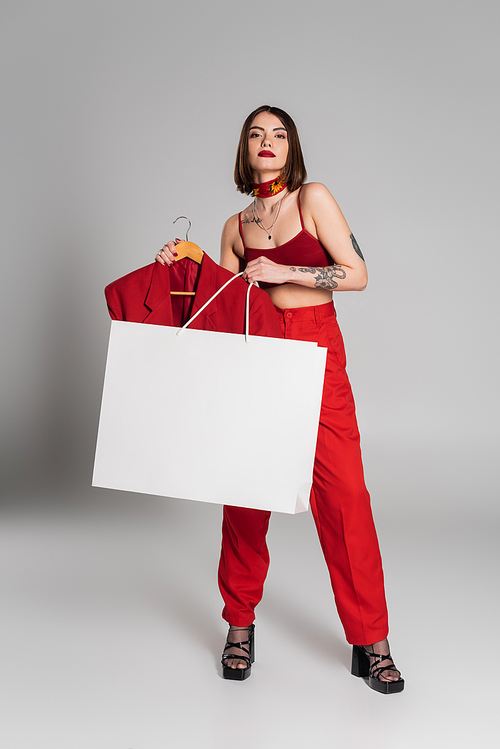 consumerism, young tattooed woman with short hair and nose piercing holding hanger with blazer and shopping bag on grey background, modern fashion trend, chic style, neck scarf, full length