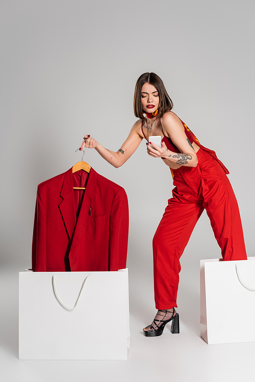 consumerism, tattooed woman with short hair and nose piercing holding hanger with blazer and using smartphone near shopping bags on grey background, modern fashion trend, full length