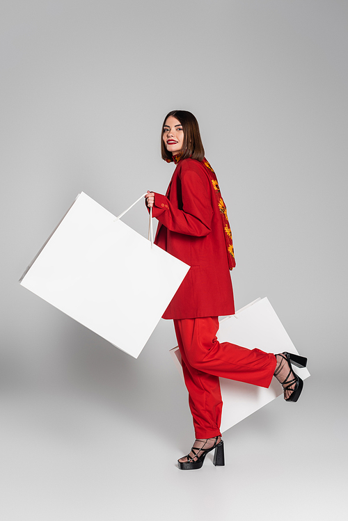 consumerism, happy woman with brunette short hair and nose piercing holding shopping bags and walking on grey background, modern fashion trend, fashionable outfit, red suit, full length