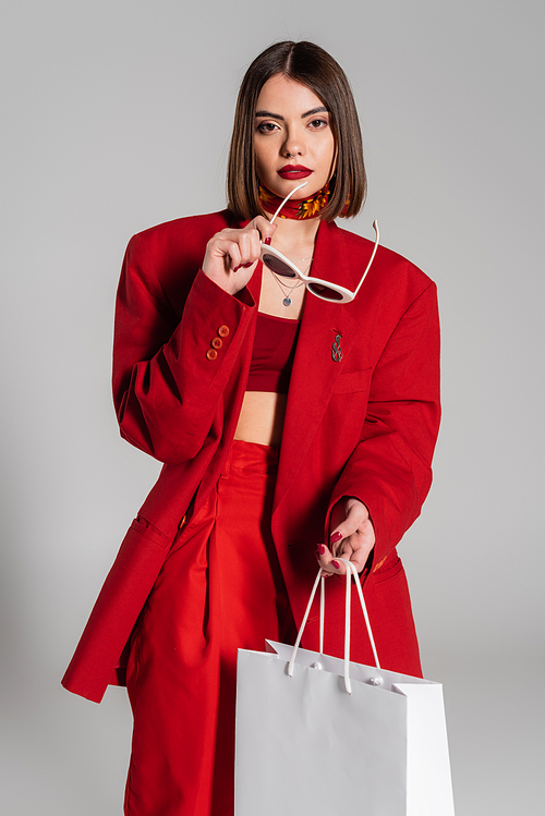 fashionable, generation z, shocked young woman with brunette short hair and nose piercing holding sunglasses and shopping bag on grey background, youth culture, red suit, consumerism