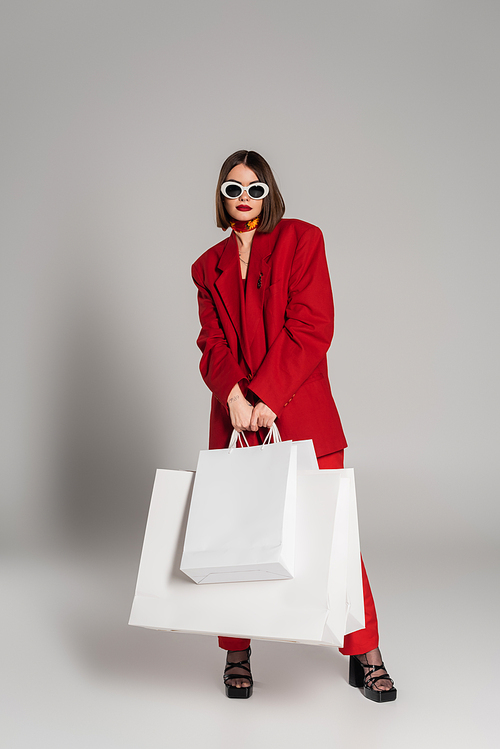 fashionable, generation z, young woman with brunette short hair and nose piercing posing in sunglasses and holding shopping bags on grey background, youth culture, red suit, consumerism