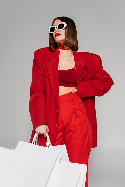 fashionable, generation z, young woman with brunette short hair and nose piercing posing in sunglasses and holding shopping bags on grey background, lady in red, consumerism