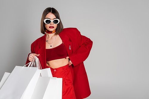 fashionable, generation z, young woman with brunette short hair and nose piercing posing in sunglasses and red suit while holding shopping bags on grey background, youth culture, consumerism