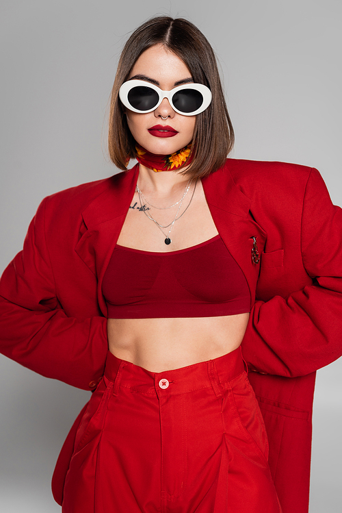 generation z, tattooed young woman with short hair and nose piercing posing in sunglasses and red suit on grey background, modern fashion, trendy outfit, chic style