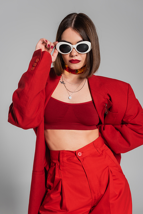 trendy outfit, generation z, tattooed young woman with brunette short hair and nose piercing posing in sunglasses and red suit on grey background, modern fashion, chic style