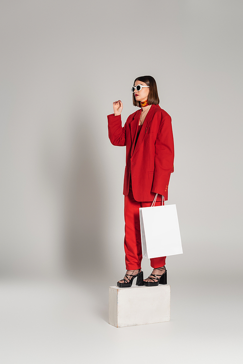 generation z, consumerism, young woman with brunette short hair and nose piercing posing in sunglasses and red suit while holding shopping bag and standing on concrete cube on grey background