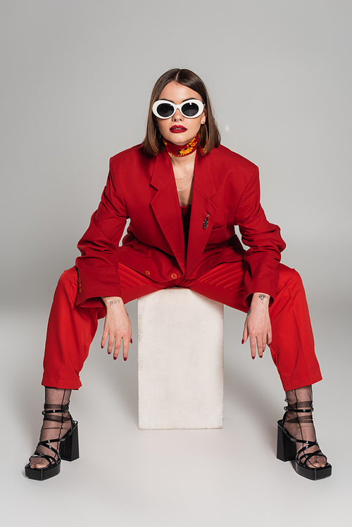 generation z, fashion model with brunette short hair and nose piercing posing in sunglasses and red suit while sitting on concrete cube on grey background, lady in red, fashionista