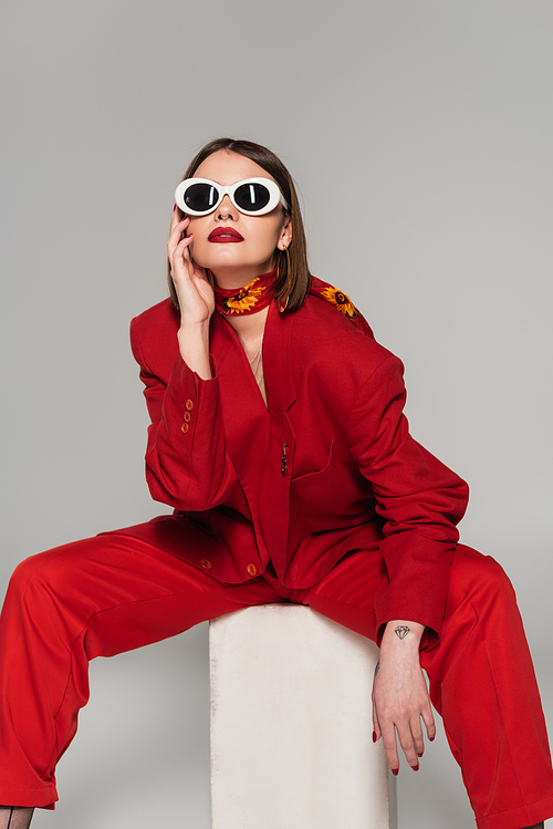 generation z, fashion model with brunette short hair and nose piercing posing in sunglasses and red suit while sitting on concrete cube on grey background, lady in red, young woman in red outfit