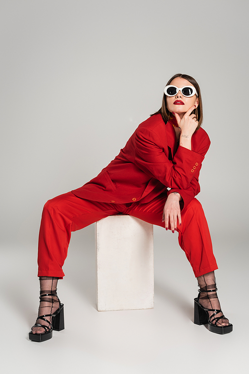 generation z, fashion model with brunette short hair and nose piercing posing in sunglasses and red suit while sitting on concrete cube on grey background, lady in red, young woman