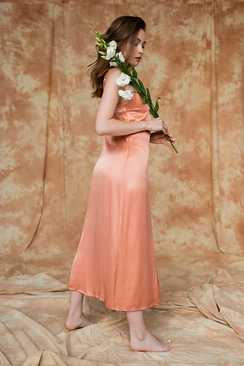 full length of barefoot and brunette and young woman in pink silk slip dress holding eustoma flowers while standing on mottled beige background, sensuality, elegance, sophistication