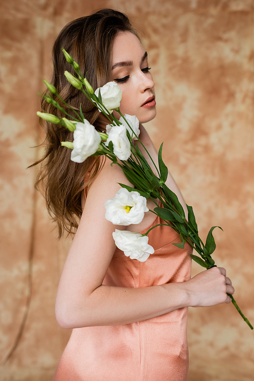 portrait of brunette and young woman in pink silk slip dress holding eustoma flowers while standing on mottled beige background, sensuality, elegance, sophistication