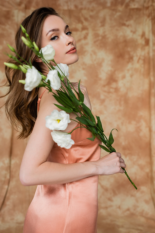 portrait of graceful, brunette and young woman in pink silk slip dress holding eustoma flowers while standing on mottled beige background, sensuality, elegance, sophistication, looking at camera