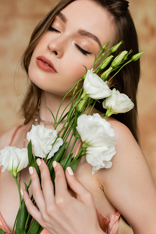 portrait of dreamy, brunette and young woman with closed eyes holding eustoma flowers while standing and posing on mottled beige background, sensuality, elegance, sophistication