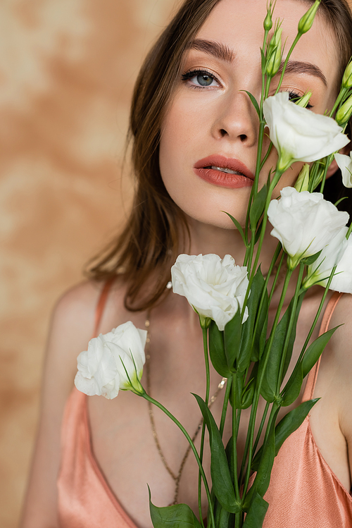 portrait of sensual and young woman holding eustoma flowers while standing and looking at camera on mottled beige background, captivating beauty, elegance, sophistication