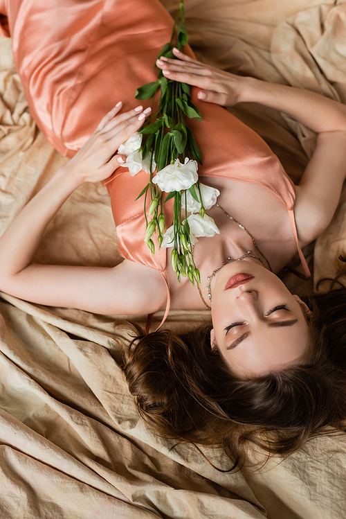 top view of young woman in pink silk slip dress lying on linen fabric with closed eyes and holding white flowers on mottled beige background, sensuality, sophistication, elegance, eustoma