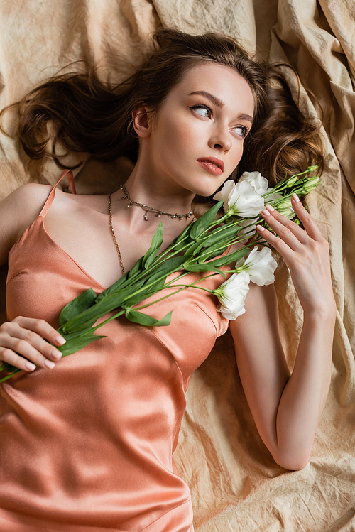 top view of attractive young woman in pink silk slip dress lying on linen fabric and holding delicate white flowers on beige background, sensuality, elegance, eustoma, looking away