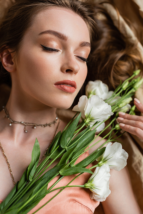 top view of attractive young woman in pink silk slip dress lying with closed eyes on linen fabric and holding white flowers on beige background, sensuality, elegance, eustoma, portrait