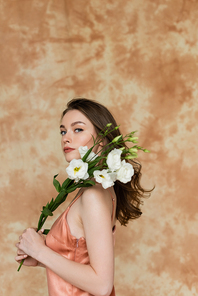 young woman with brunette hair posing in pink and silk slip dress and holding white eustoma flowers on mottled beige background, sensuality, sophistication, elegance, looking at camera